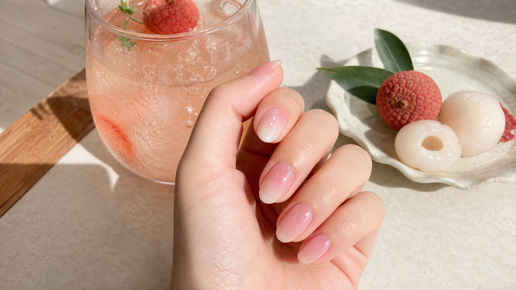 Lychee-inspired sheer pink and milky white gel nails beside a chilled lychee drink and fresh fruit